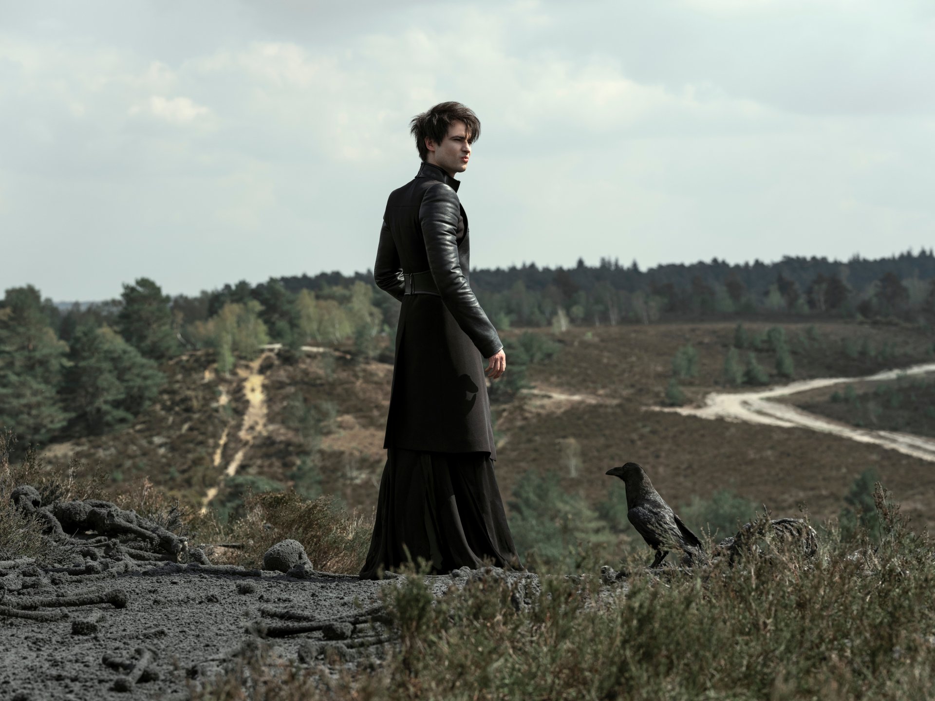 2K Quad HD PC desktop wallpaper: moody The Sandman TV show scene — a dark-clad figure on a windswept hillside with a raven, winding dirt path and distant treeline.