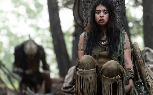 Forest-set 2K Quad HD PC desktop wallpaper: young warrior woman, dirt-streaked face and fringe buckskin, tense with a Predator figure behind — scene from the movie Prey (2022).
