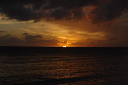HD photography of a vibrant sunset over the ocean, with the sun partially visible on the horizon and dramatic clouds casting warm hues across the sky.