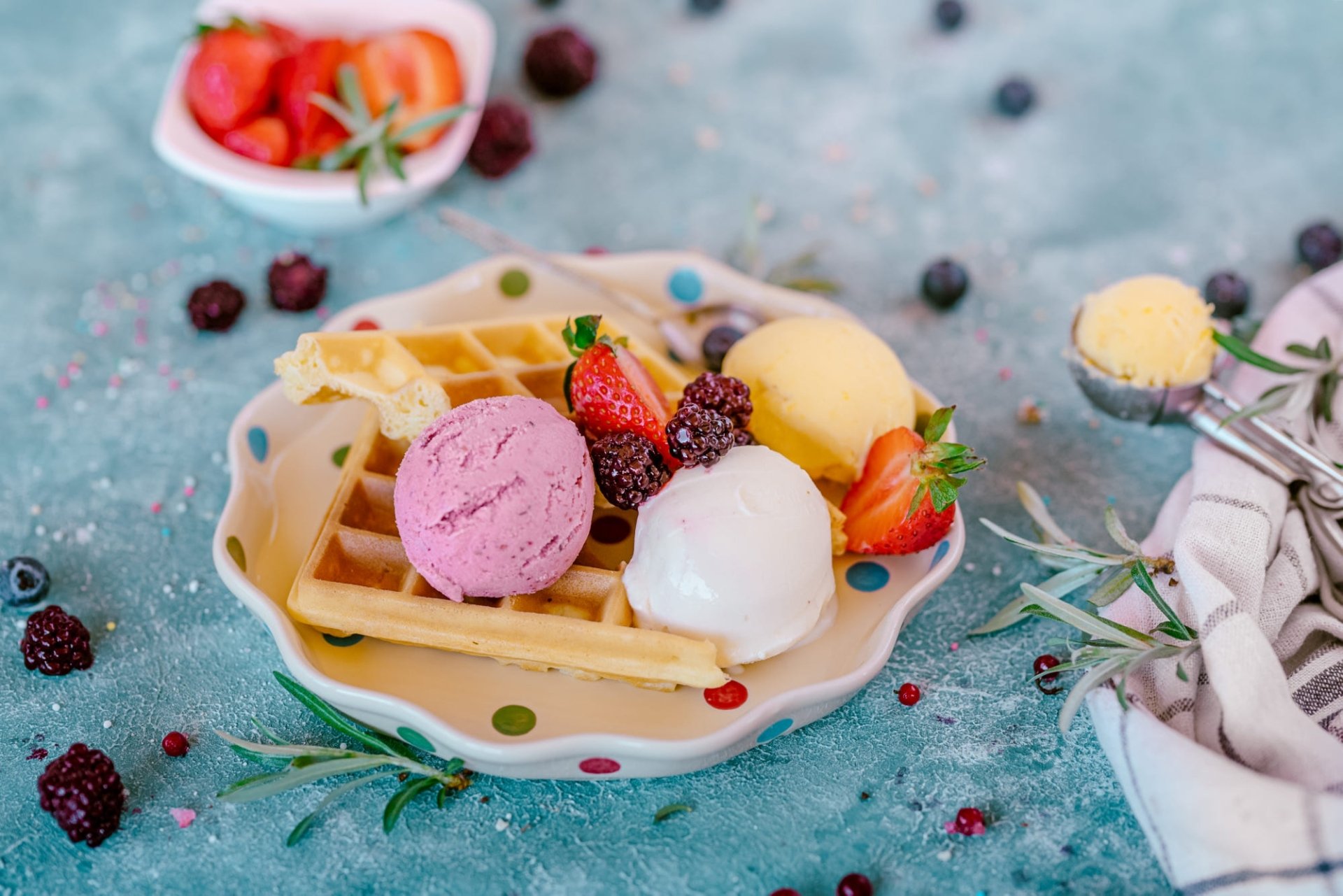 HD PC desktop wallpaper and background: colorful food scene of a waffle topped with pink, white, and yellow ice cream scoops, strawberries and mixed berries on a textured teal surface.