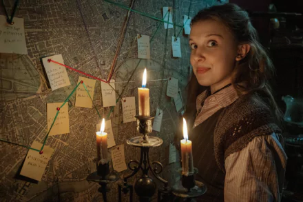 HD PC desktop wallpaper: candlelit cinematic scene from Enola Holmes 2 — a young woman inspects a wall map pinned with notes, lit by a trio of burning candles.