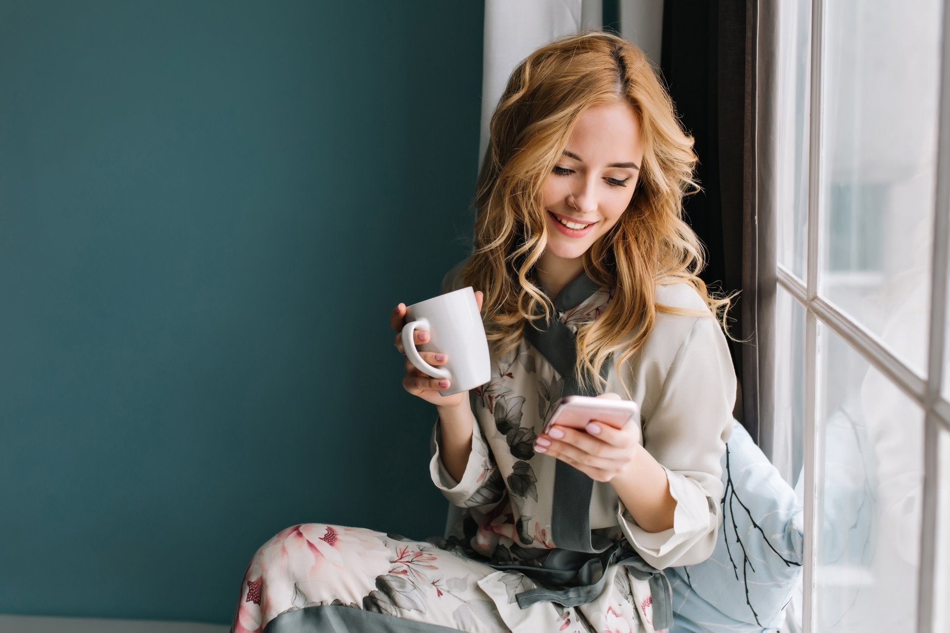 A smiling woman holding a white mug and looking at her phone, captured in a bright, cozy moment. 4K Ultra HD PC desktop wallpaper and background.