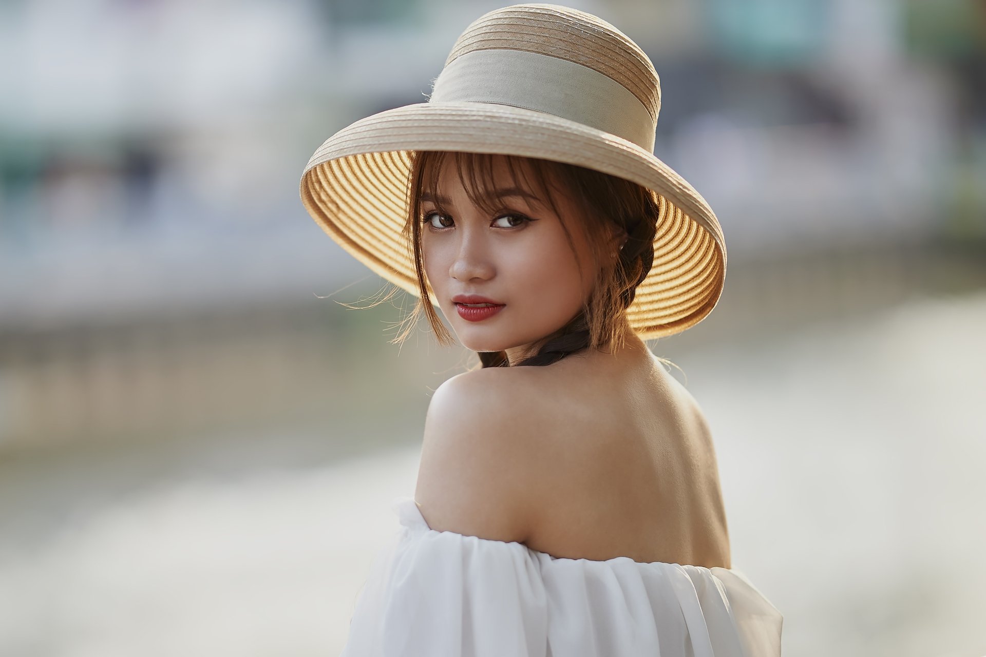 5K Ultra HD PC desktop wallpaper/background of a woman in an off-shoulder white top and straw sunhat, glancing over her shoulder against a soft bokeh waterfront backdrop.