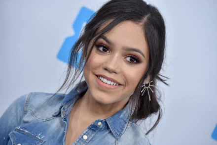 HD PC desktop wallpaper featuring celebrity Jenna Ortega smiling with natural makeup and wearing a denim shirt and star-shaped earrings.