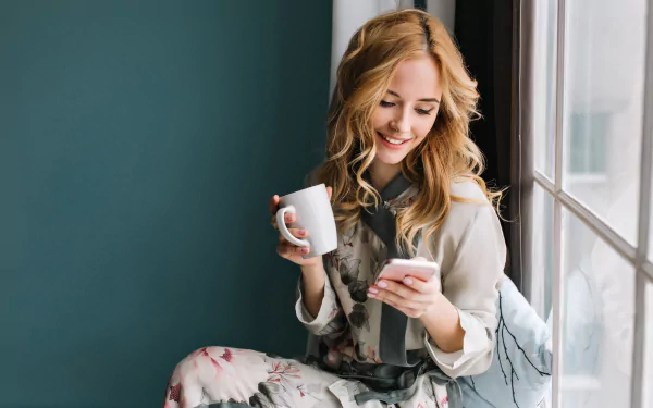 A smiling woman holding a white mug and looking at her phone, captured in a bright, cozy moment. 4K Ultra HD PC desktop wallpaper and background.