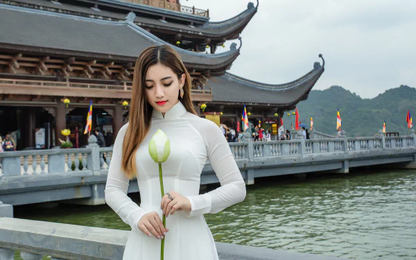 Asian woman in traditional white dress holding a lotus flower by a temple and water, captured in 4K Ultra HD for a PC desktop wallpaper.