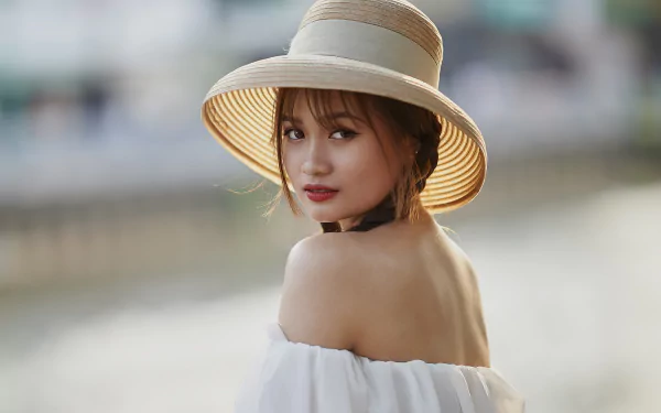 5K Ultra HD PC desktop wallpaper/background of a woman in an off-shoulder white top and straw sunhat, glancing over her shoulder against a soft bokeh waterfront backdrop.