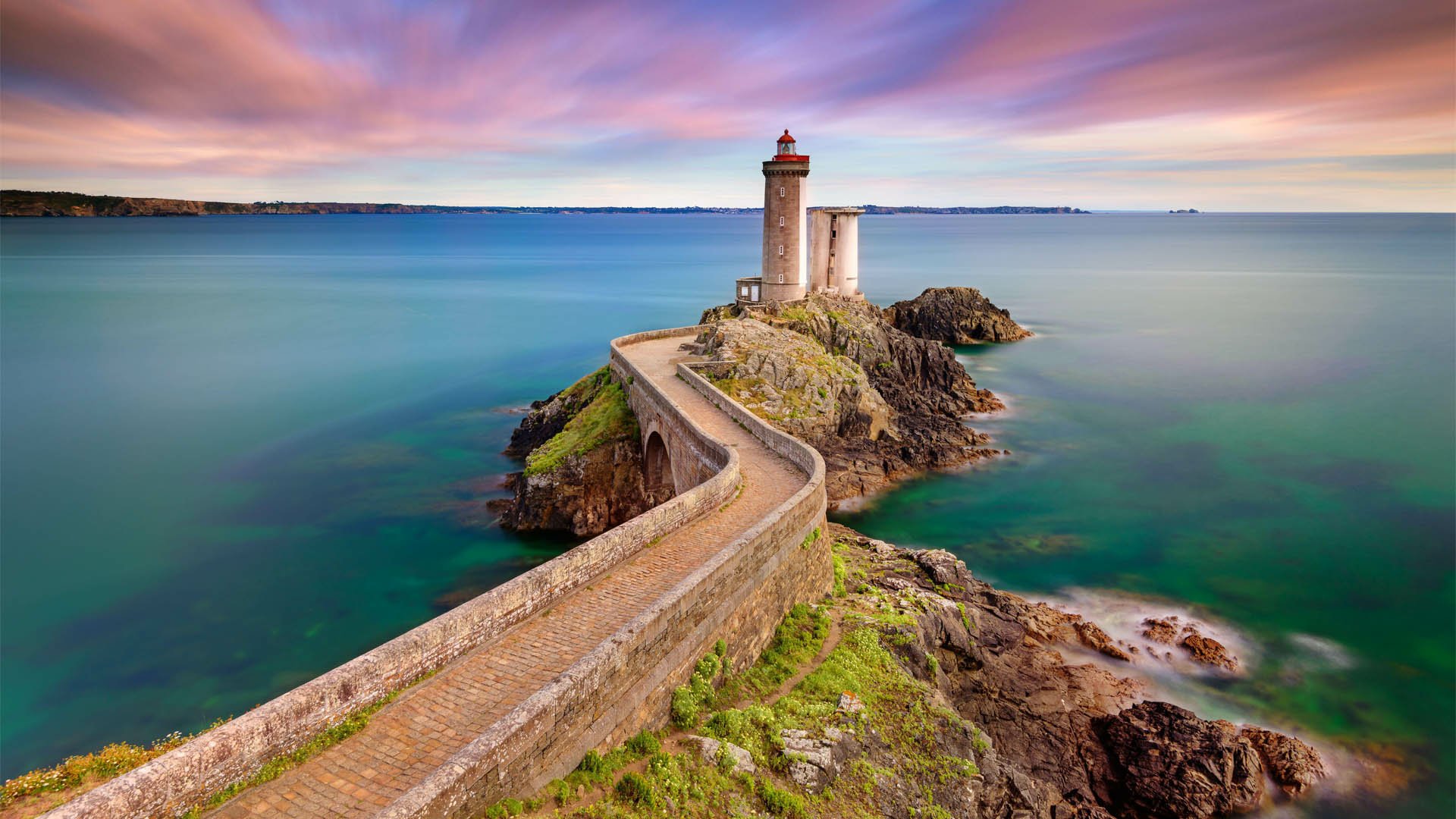 HD desktop wallpaper of a man-made lighthouse on a rocky coastline with a winding walkway under a colorful sunset sky.