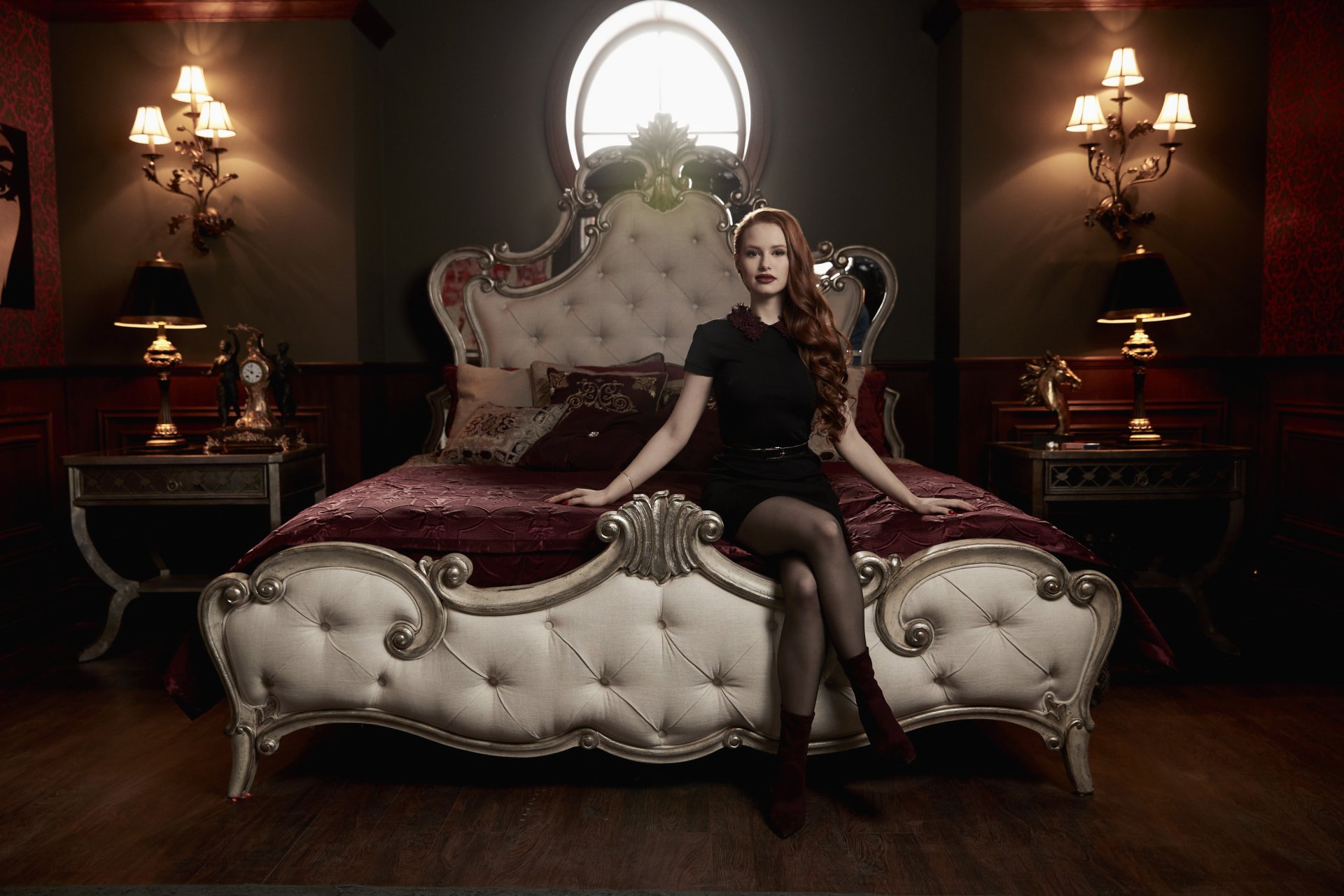 Riverdale TV show 2K Quad HD PC desktop wallpaper: red‑haired woman in black dress sits on an ornate tufted bed in a dim, candlelit, vintage bedroom.