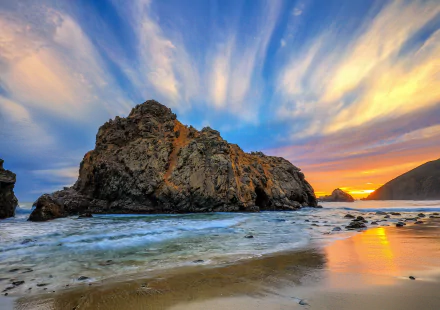 HD PC desktop wallpaper background: nature scene of a rugged rock formation on a sandy beach at sunset, dramatic streaked clouds and colorful sky reflected on wet sand.