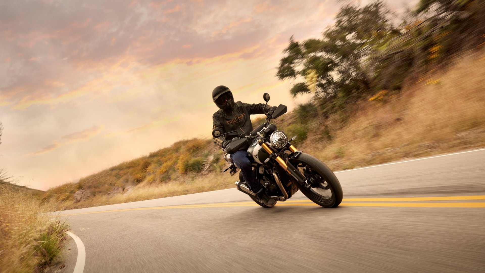 HD wallpaper featuring a rider on a Triumph Scrambler 400 X motorcycle cruising along a winding road with a dynamic, blurred background to convey speed and motion.