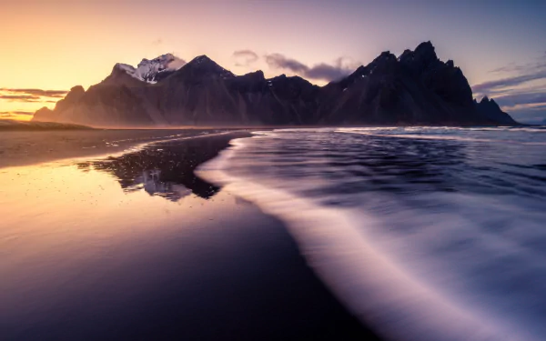 A serene 4K Ultra HD nature scene of Vestrahorn mountains at sunset with reflective black sand and gentle ocean waves.