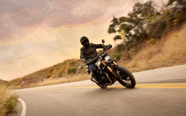 HD wallpaper featuring a rider on a Triumph Scrambler 400 X motorcycle cruising along a winding road with a dynamic, blurred background to convey speed and motion.