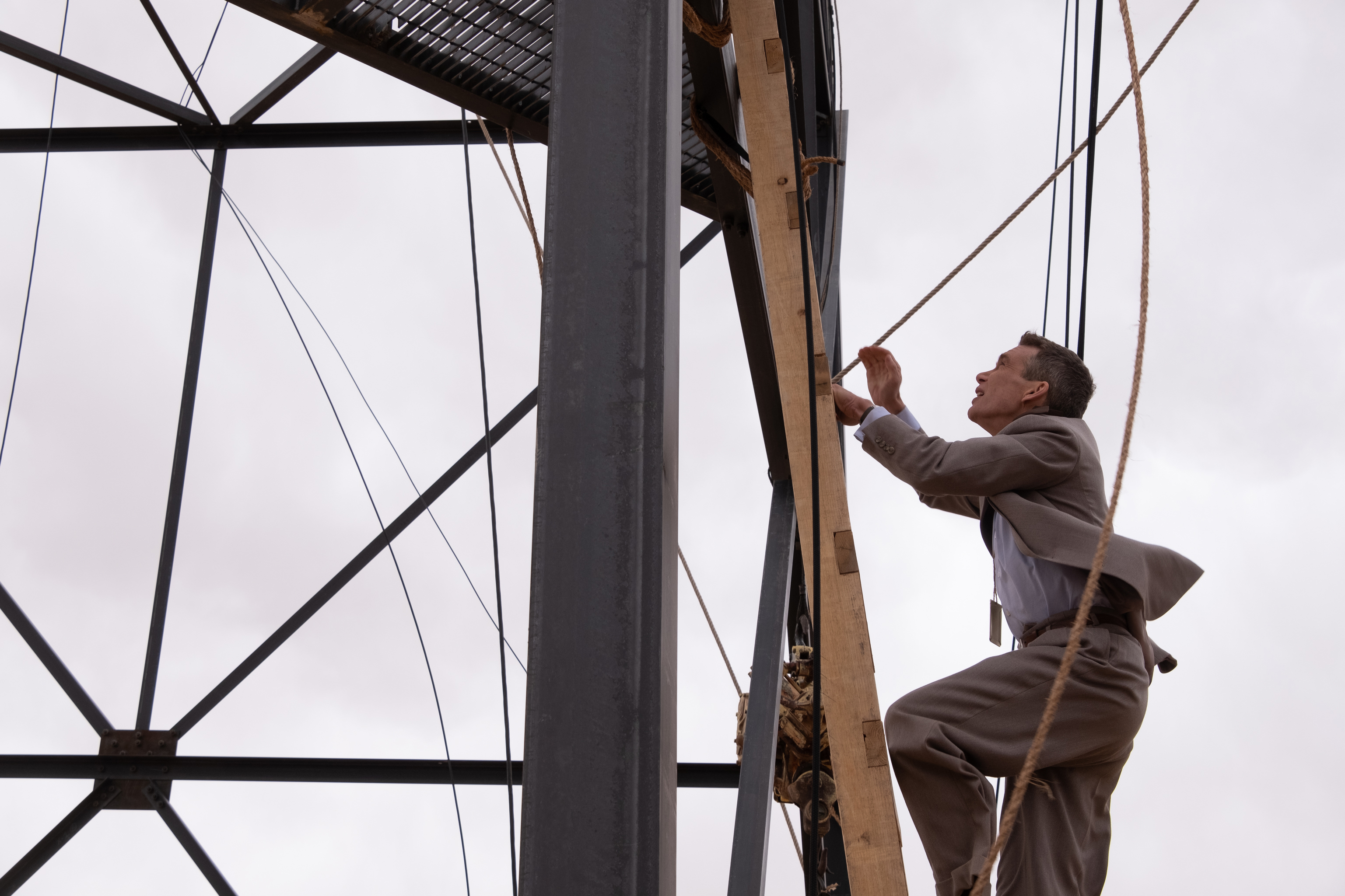 HD wallpaper of a man in a suit climbing a ladder on a scaffolding structure under a cloudy sky, tagged with Oppenheimer.