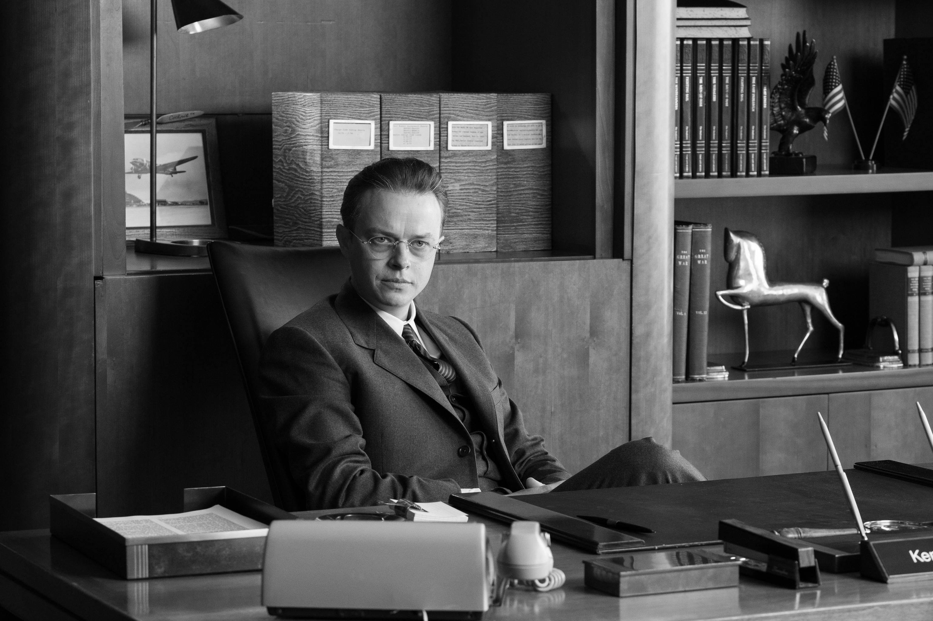 Black and white HD wallpaper featuring a poised man in a suit sitting at a desk, evoking the theme of the film Oppenheimer, for desktop background use.