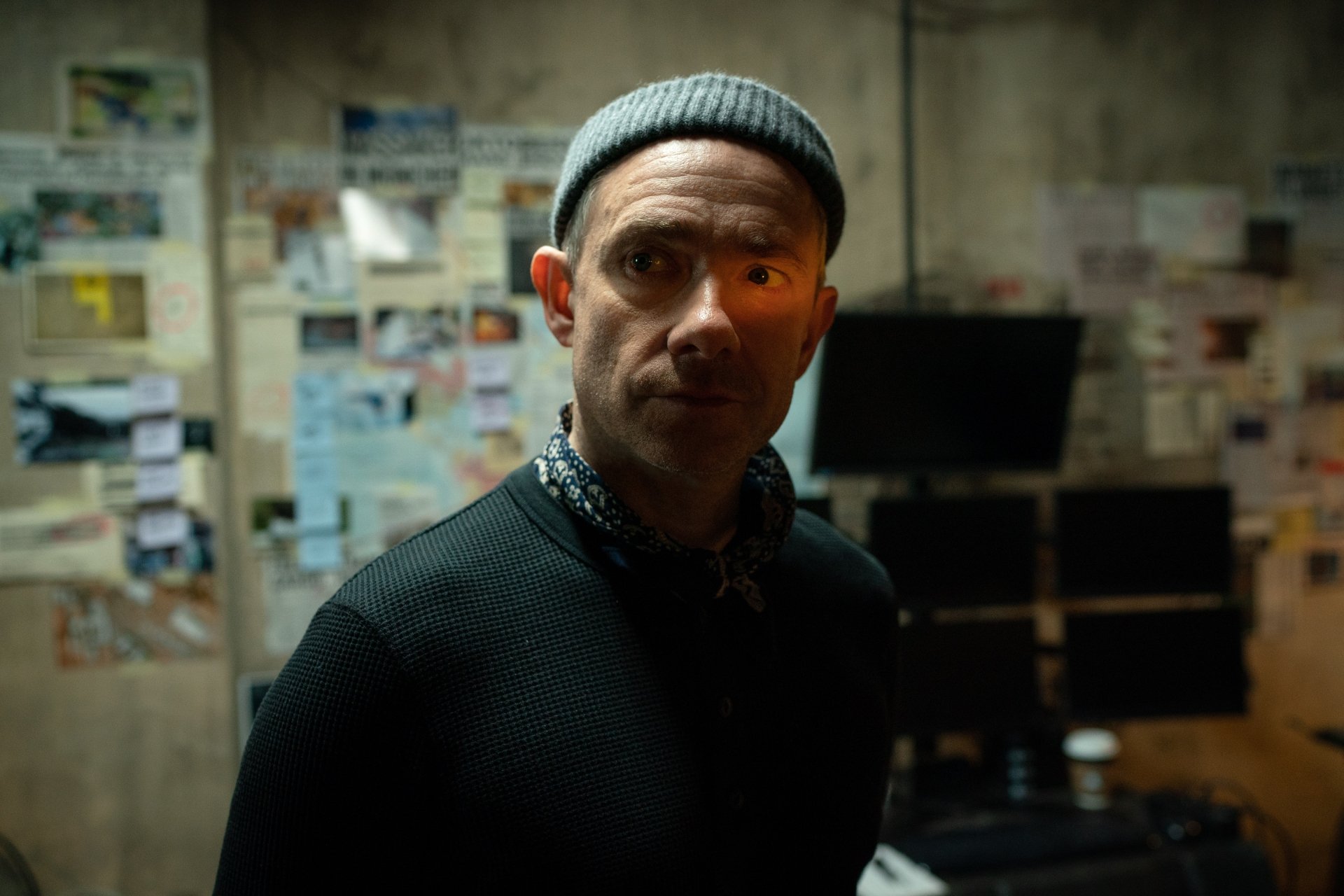 Man standing in a dimly lit room with a wall covered in various papers and notes, with a serious expression, in a Secret Invasion themed HD desktop wallpaper and background.