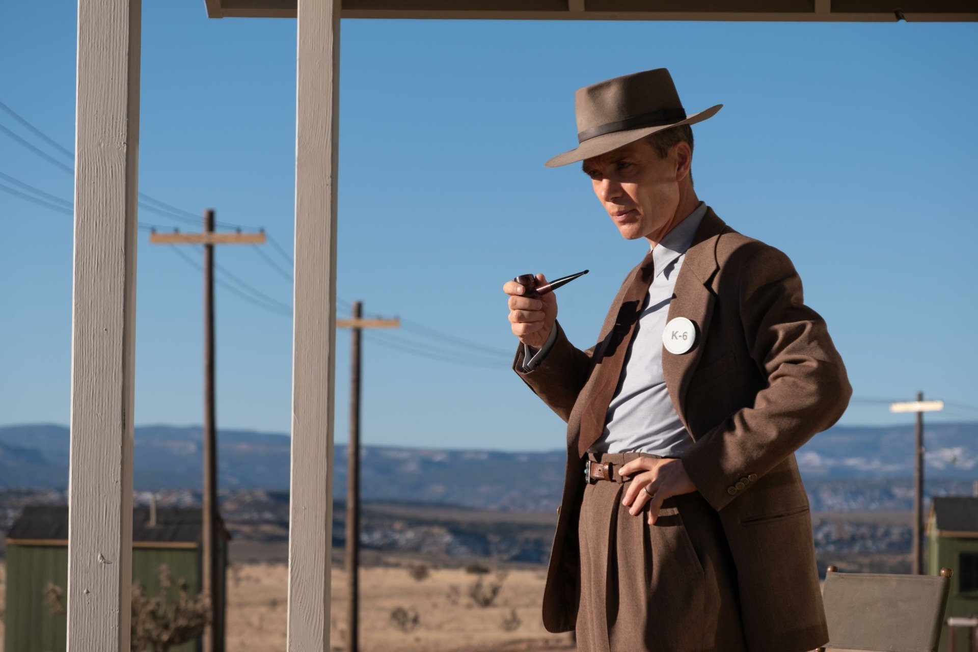 HD desktop wallpaper featuring a man styled in early to mid-20th century attire with a fedora and suit, holding a pipe, emulating the historical figure Oppenheimer, in a desert setting with power lines and expansive sky in the background.