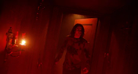 A character steps through a dimly lit hallway featuring a glowing red door, evoking the eerie atmosphere of *Insidious: The Red Door*. This image serves as a striking HD wallpaper.