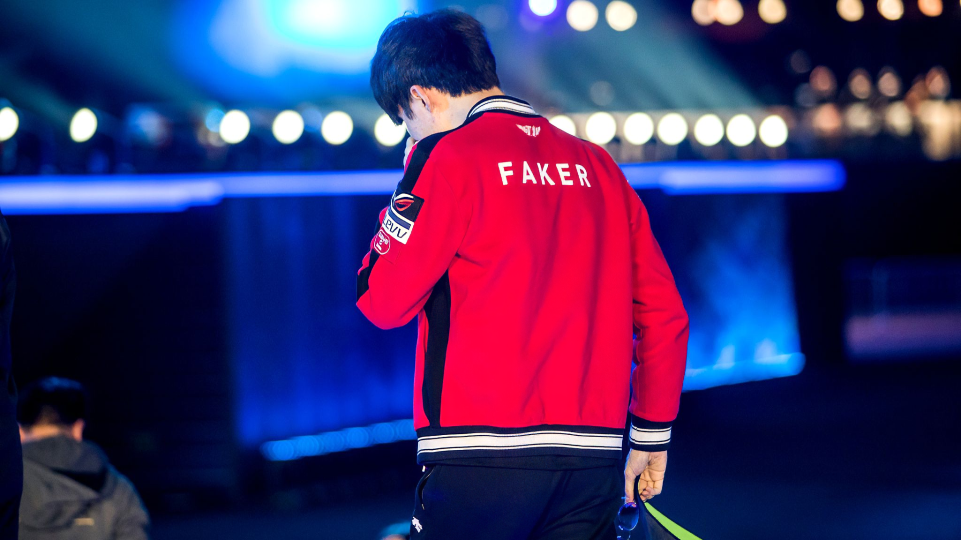 HD wallpaper featuring T1's Faker, a prominent League of Legends player, in his team jacket at a gaming event, walking on stage with a focused demeanor.