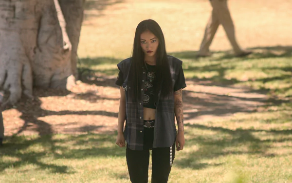 A character from the TV show The Brothers Sun stands confidently in a sunlit park, wearing a grunge-inspired outfit, surrounded by lush greenery. HD desktop wallpaper background.