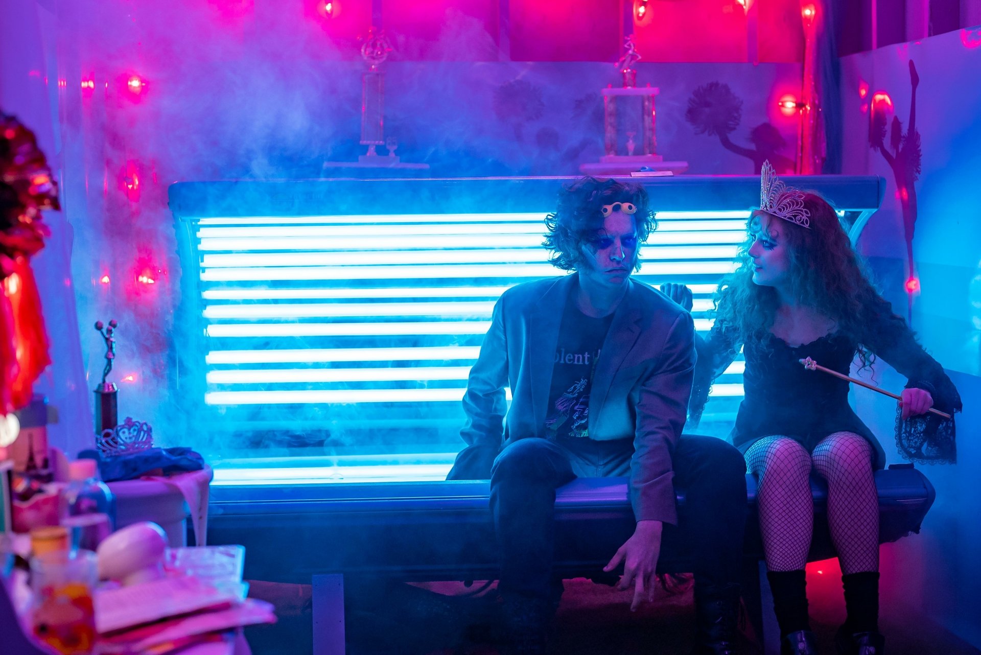 A vibrant scene from the movie Lisa Frankenstein, featuring two characters sitting on a glowing blue bench amidst colorful smoke and eclectic decor, designed as an HD desktop wallpaper.