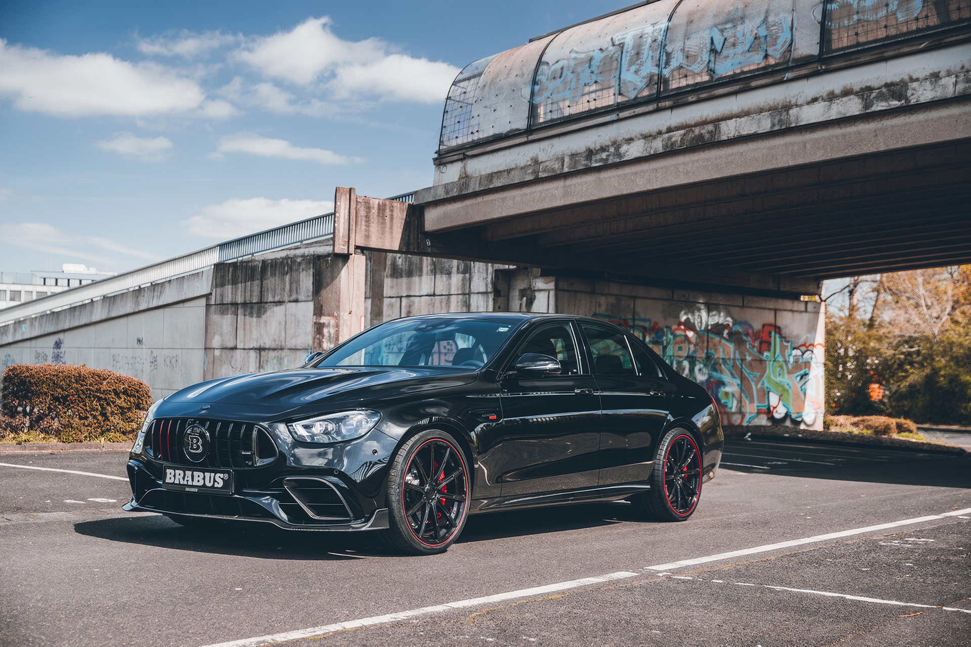 A sleek black Brabus 800 E63 parked under an overpass, showcasing its aggressive design and sporty features, set against a backdrop of urban art and a clear blue sky.
