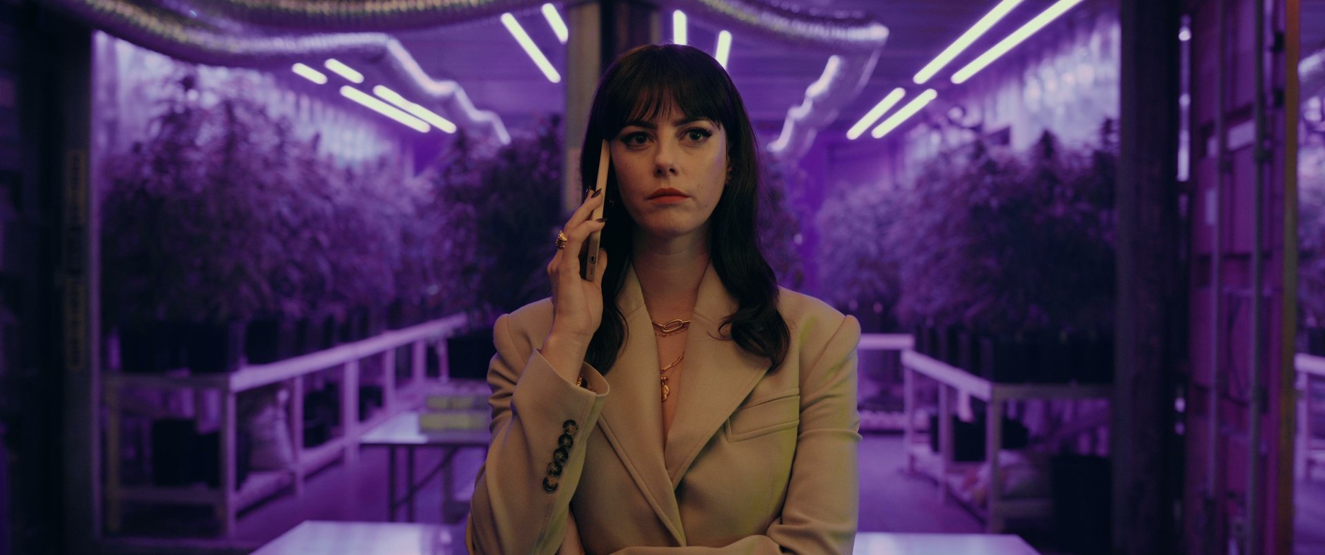 Actress in The Gentlemen (2024) TV show depicted in an HD desktop wallpaper, featuring Kaya Scodelario in a beige suit on a phone call, with a neon-lit setting in the background.