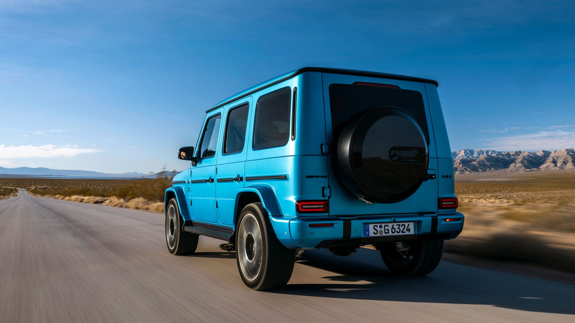 Blue Mercedes-AMG G63 driving on a desert road with mountains in the background, HD desktop wallpaper.