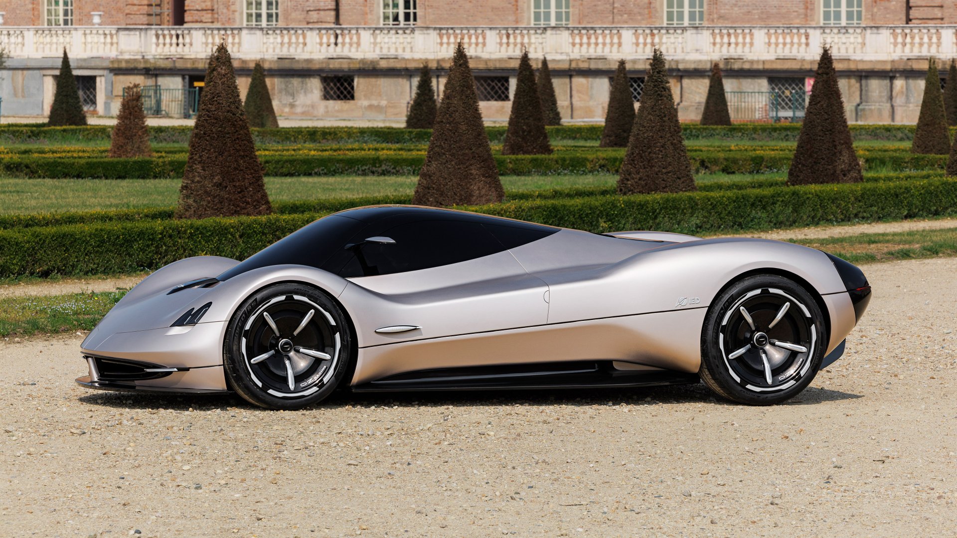 A sleek Pagani Alisea sports car parked on a gravel surface with manicured trees and a classic building in the background, suitable as a desktop wallpaper.
