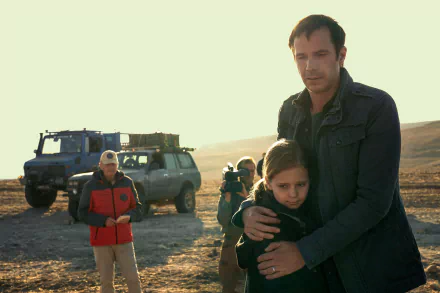 HD desktop wallpaper featuring a scene from the TV show Constellation (2024) with a man embracing a child in a desolate landscape, a vehicle, and film crew in the background.