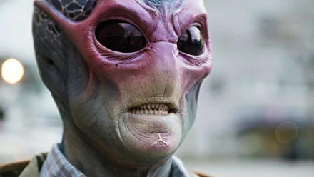 A close-up of an alien character from the TV show Resident Alien, featuring distinct purple and green skin, large black eyes, and an expressive facial structure, set against a blurred background.