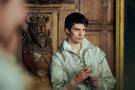 Nicholas Galitzine portraying a character from the TV show Mary & George in a high-definition wallpaper, featuring a close-up in vintage attire set against a historical wooden backdrop.