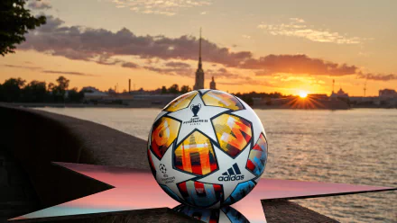 A vibrant UEFA Champions League ball rests on a star-shaped base by a river, with a stunning sunset and city silhouette in the background, creating an inspiring HD desktop wallpaper.