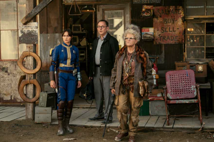 HD wallpaper featuring three characters from the Fallout TV show standing in a rustic, post-apocalyptic setting with aged props and signs indicating trade and commerce.