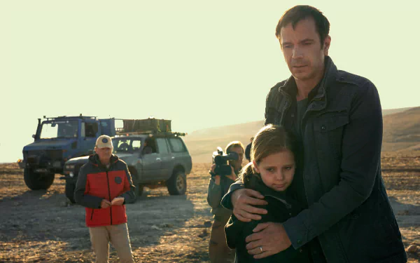 HD desktop wallpaper featuring a scene from the TV show Constellation (2024) with a man embracing a child in a desolate landscape, a vehicle, and film crew in the background.