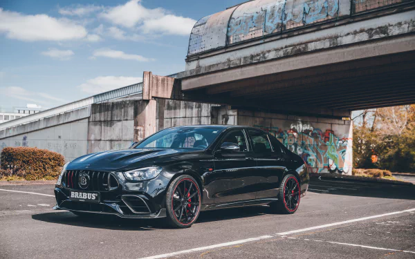 A sleek black Brabus 800 E63 parked under an overpass, showcasing its aggressive design and sporty features, set against a backdrop of urban art and a clear blue sky.