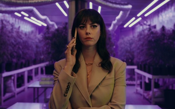 Actress in The Gentlemen (2024) TV show depicted in an HD desktop wallpaper, featuring Kaya Scodelario in a beige suit on a phone call, with a neon-lit setting in the background.