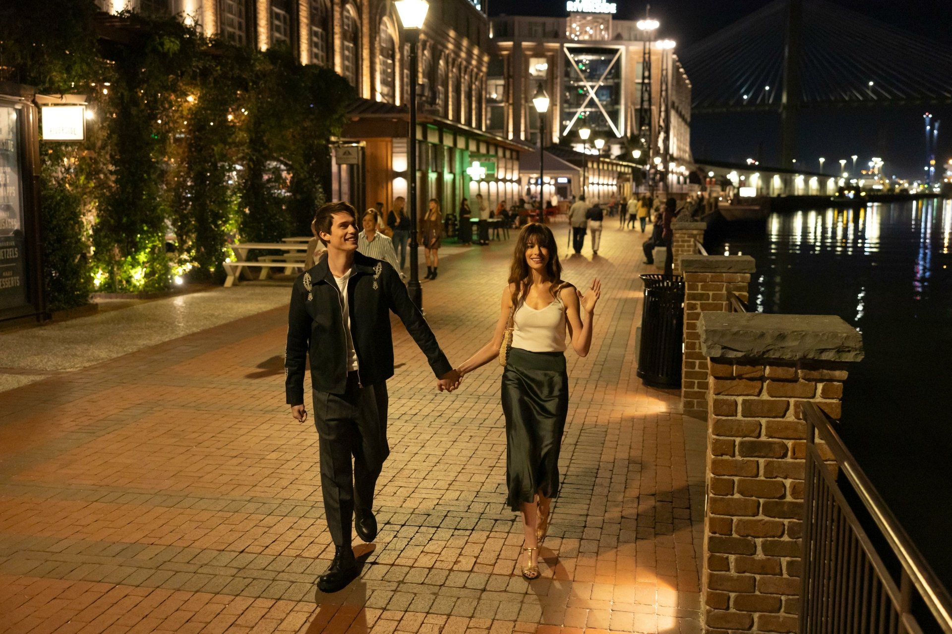 HD desktop wallpaper from The Idea of You featuring Anne Hathaway and Nicholas Galitzine walking hand in hand by a riverwalk at night.
