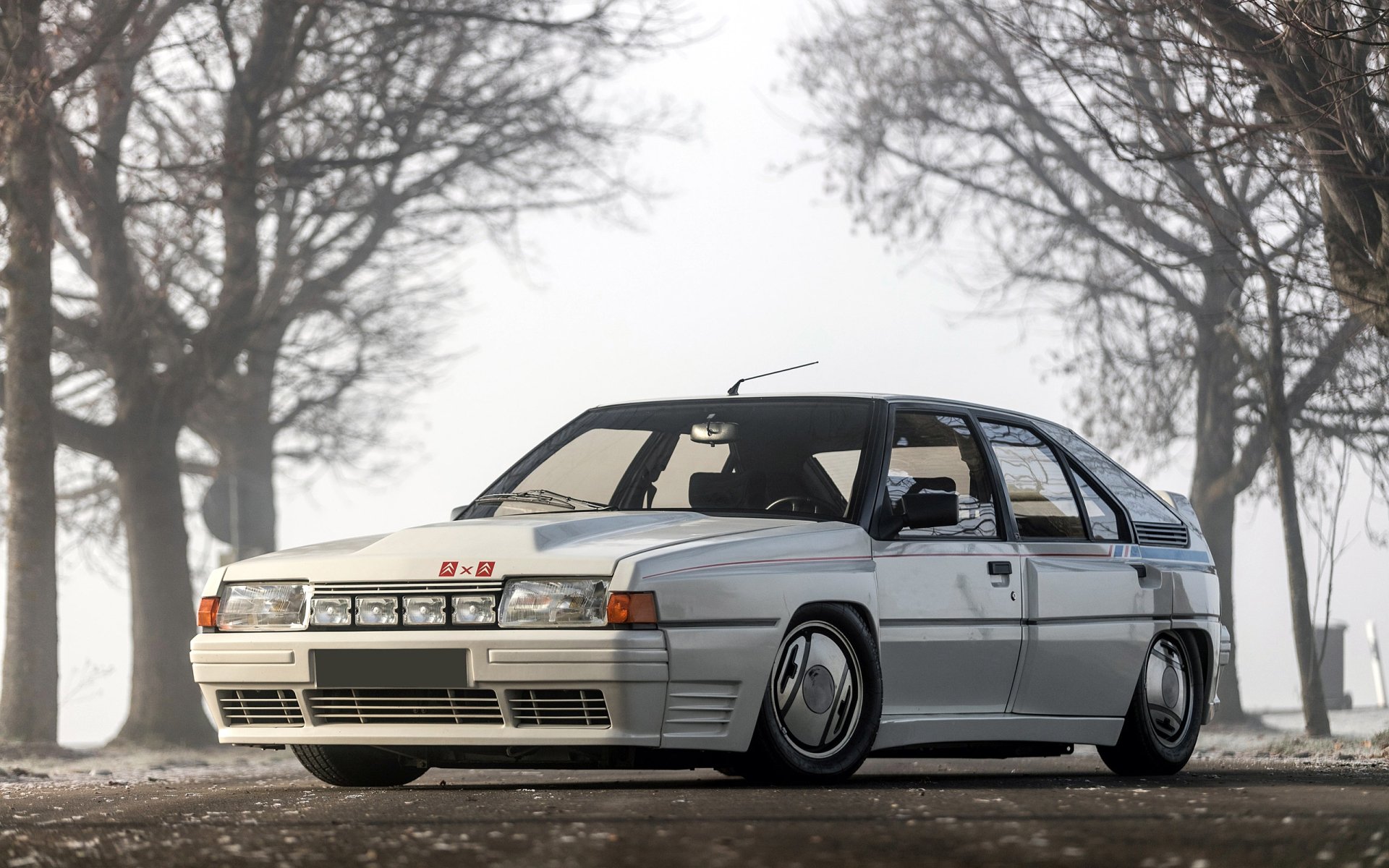 HD image of a classic white Citroën BX 4TC car, set as a desktop wallpaper. The vehicle is parked on a misty road with bare trees in the background.