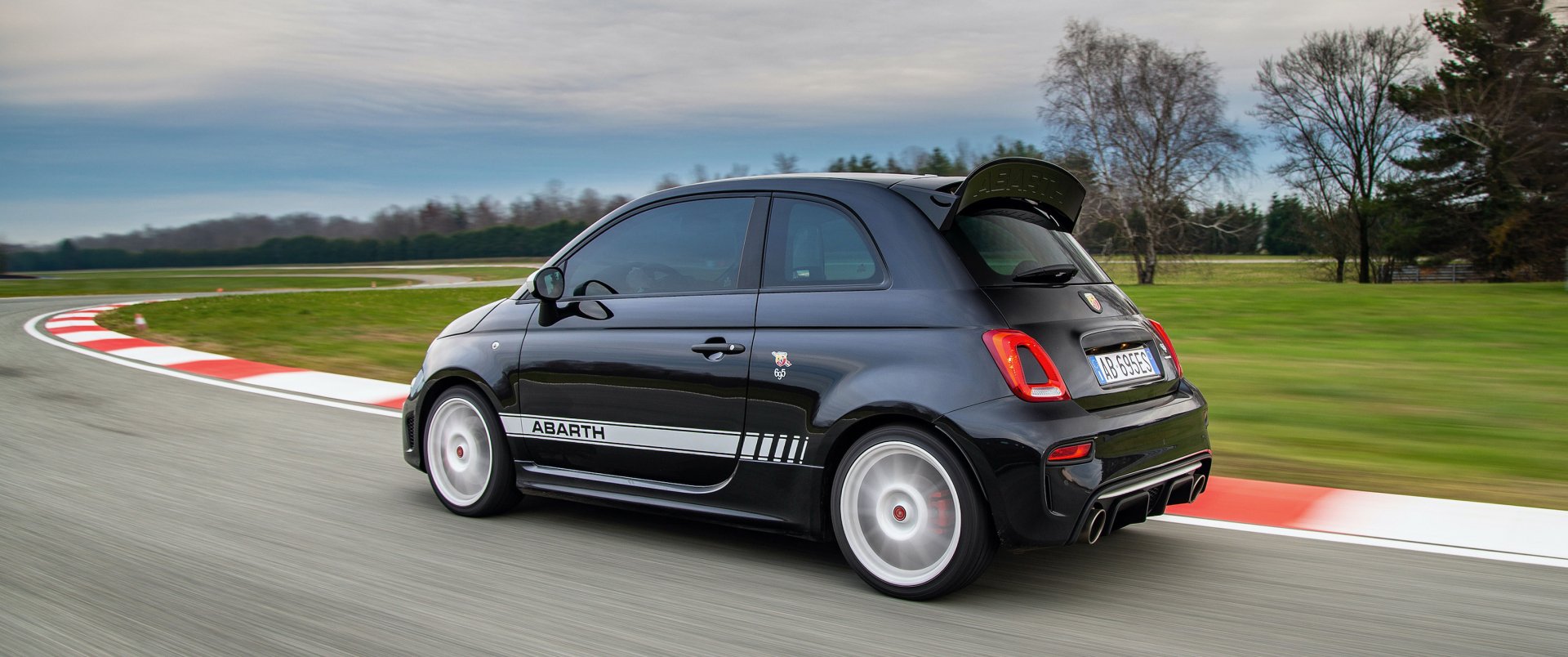 HD desktop wallpaper featuring a sleek black Fiat Abarth 695 Esseesse speeding on a racetrack, showcasing its sporty design against a blurred background of green fields and trees.