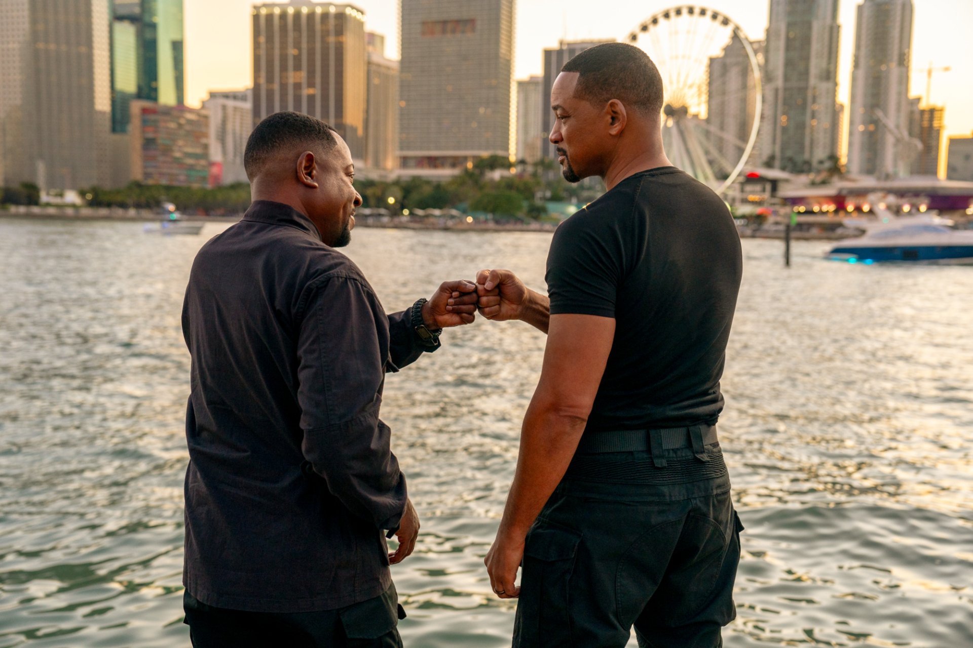 HD wallpaper from Bad Boys: Ride or Die featuring Will Smith and Martin Lawrence fist-bumping at a waterfront with a cityscape in the background.