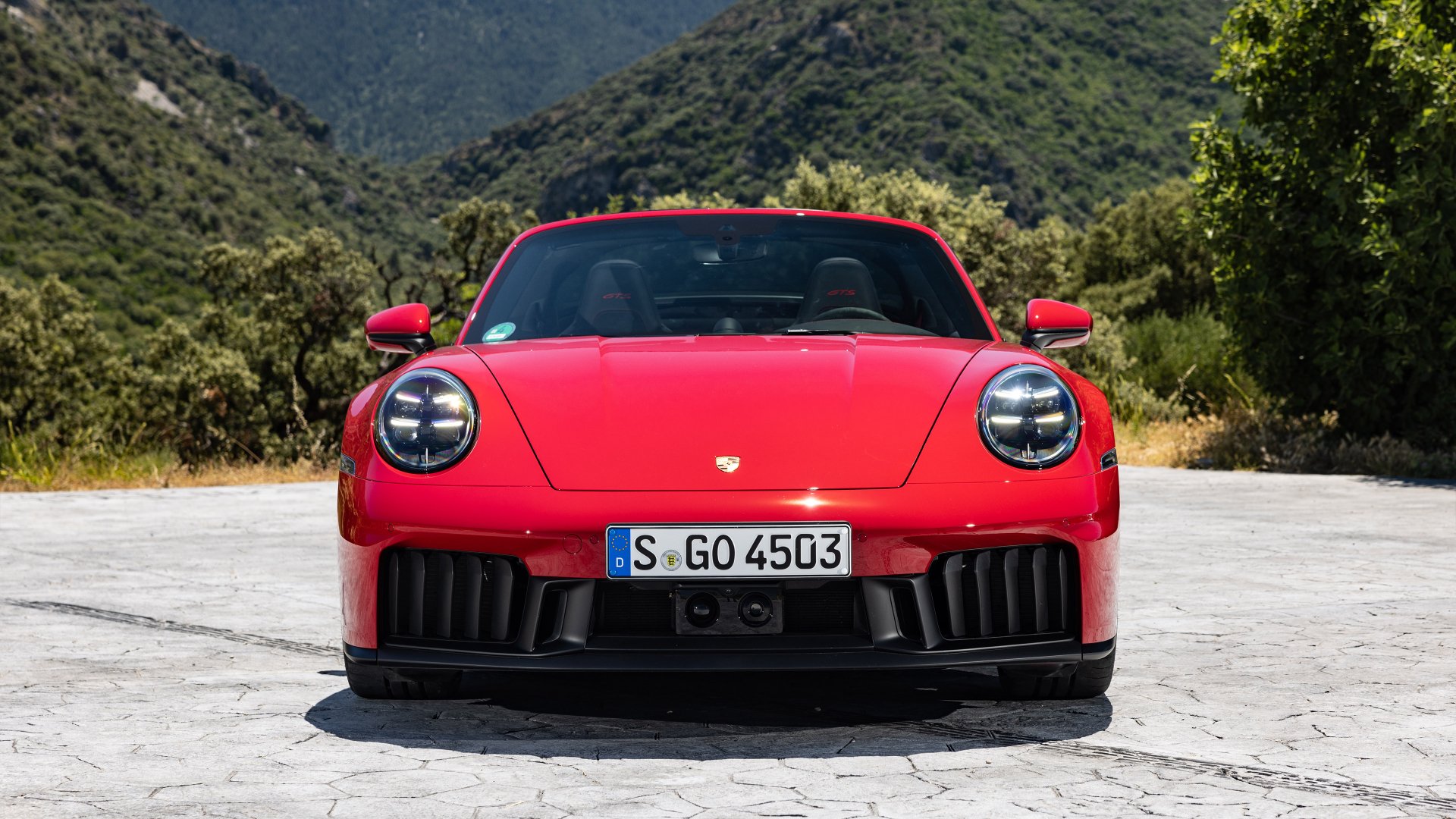 A HD desktop wallpaper featuring the front view of a striking red Porsche 911 Targa 4 GTS, set against a backdrop of mountains and greenery.