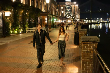 HD desktop wallpaper from The Idea of You featuring Anne Hathaway and Nicholas Galitzine walking hand in hand by a riverwalk at night.