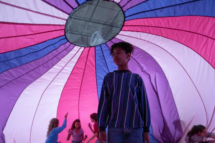 HD desktop wallpaper featuring a scene from the movie I Saw the TV Glow, showing a person standing inside a vibrant, multicolored parachute with others visible in the background.