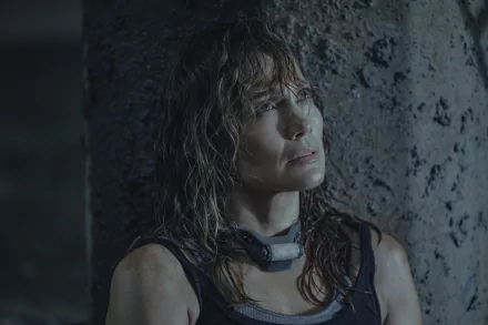 A HD desktop wallpaper featuring a scene from Atlas (2024), showing a determined woman with wet hair leaning against a weathered stone wall.