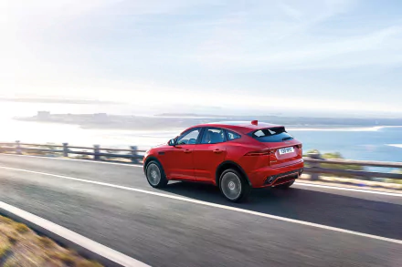 HD desktop wallpaper featuring a red Jaguar E-Pace driving along a scenic coastline road, with the ocean and bright sky in the background.