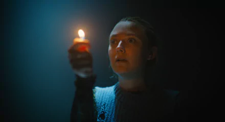 HD wallpaper of Dakota Fanning in The Watchers (2024), holding a lit candle in a dark setting, illuminating her face with a soft glow.