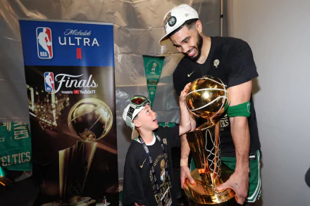 NBA player Jayson Tatum of the Boston Celtics joyfully interacts with a young fan while holding the championship trophy. The image captures a memorable moment from the NBA Finals.