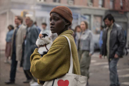 HD desktop wallpaper featuring Lupita Nyong'o in a scene from A Quiet Place: Day One. She is wearing a beanie and holding a stuffed animal on a busy urban street.