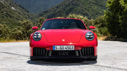A HD desktop wallpaper featuring the front view of a striking red Porsche 911 Targa 4 GTS, set against a backdrop of mountains and greenery.